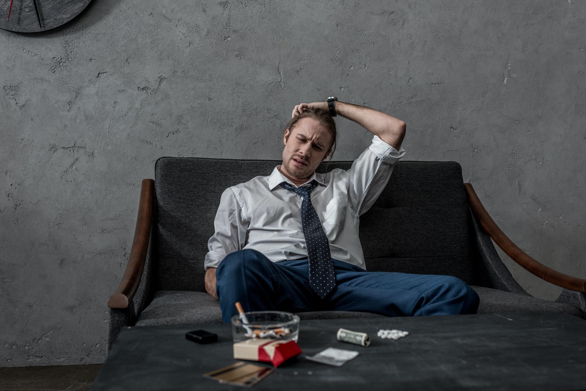 depressed businessman struggling with substance abuse sitting on couch in front of table with drugs