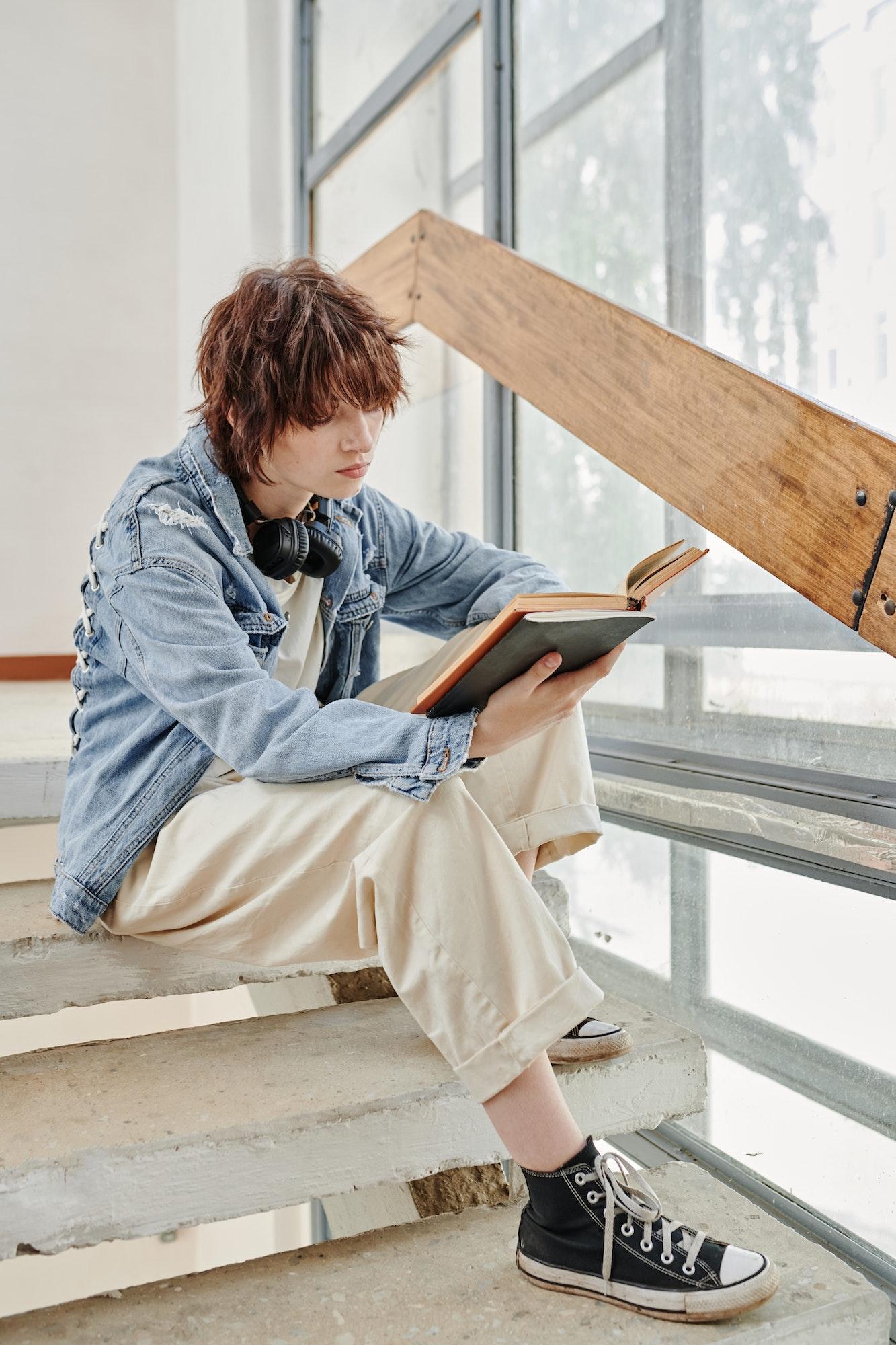 Serious youthful girl in casualwear sitting on staircase and reading book